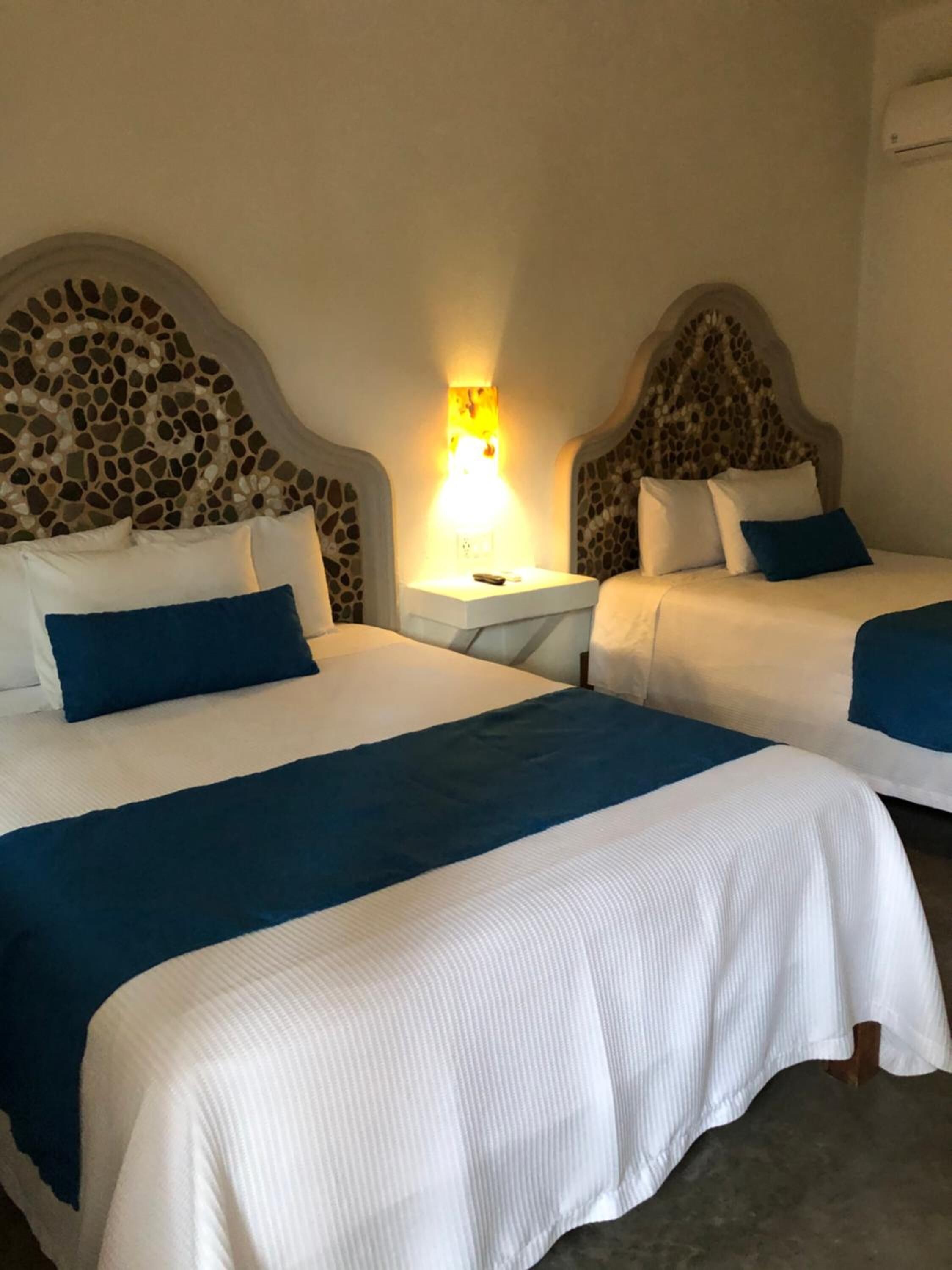 Habitación de hotel con dos camas, cada una con ropa de cama blanca y detalles en azul, cabecera con estampado de piedra y mesita de noche con lámpara.