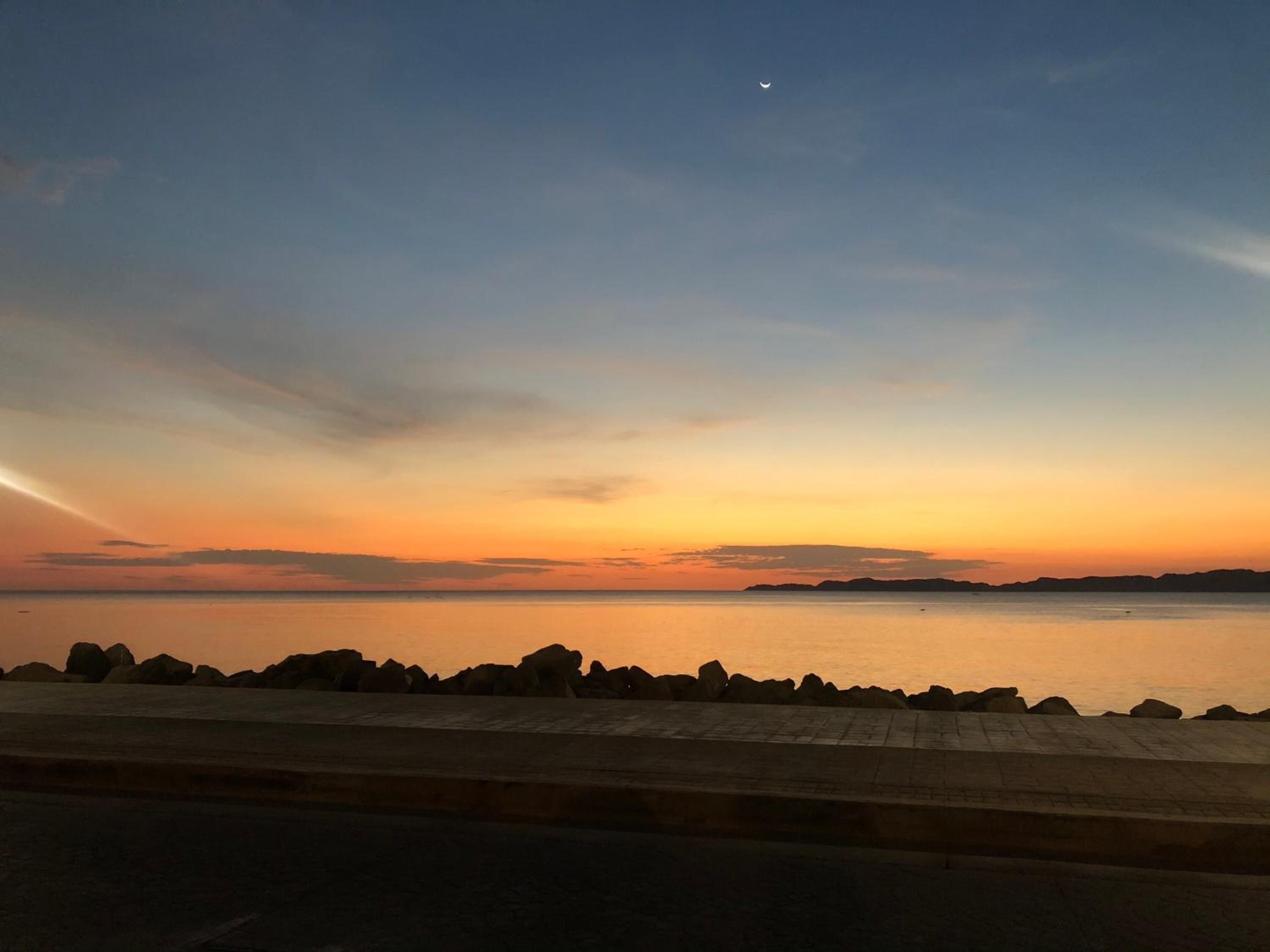 Un coucher de soleil sur une mer calme, avec une côte rocheuse et une terre lointaine à l’horizon.