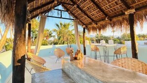 A covered outdoor seating area with a thatched roof, concrete tables, and wicker chairs. - Paraiso Azul Hotel (Loreto)