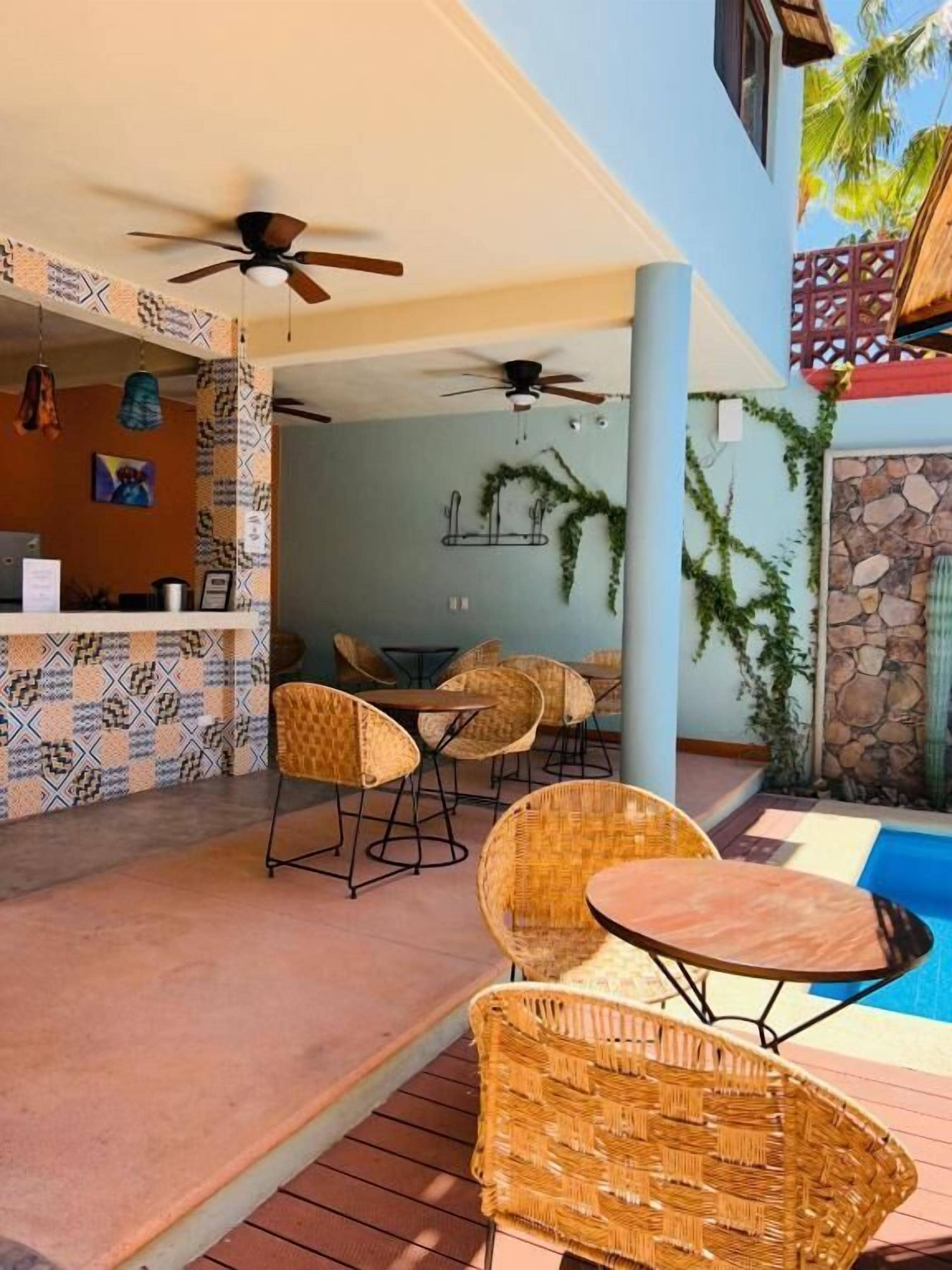 Un espace aménagé au bord de la piscine avec des fauteuils en osier et une table, un coin bar avec des carreaux décoratifs et un ventilateur de plafond.