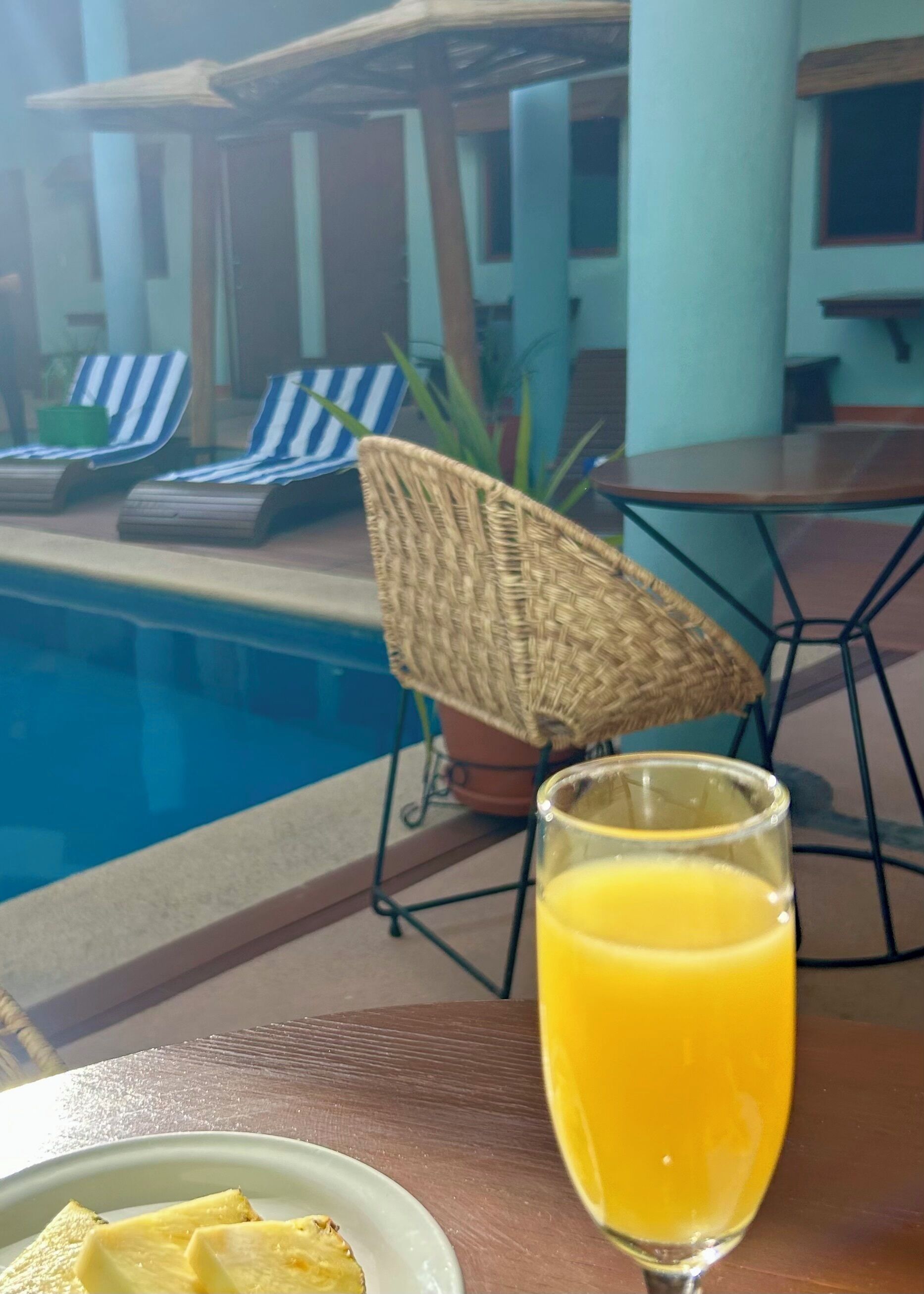 Un espace aménagé au bord de la piscine, avec des chaises longues, une table sur laquelle se trouve un verre de jus d’orange et un parasol en osier.
