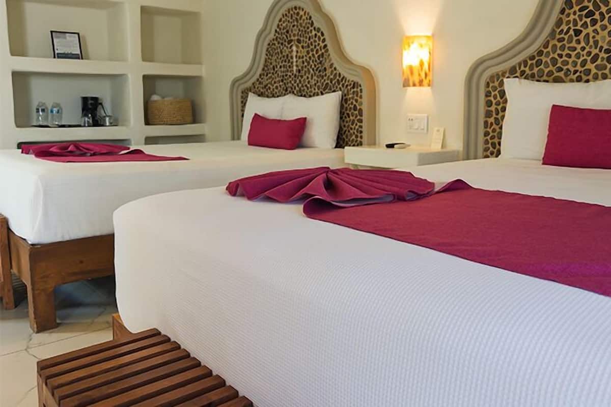 A hotel room with two beds, a headboard with a unique pattern, a nightstand with a lamp, and a shelf with toiletries.