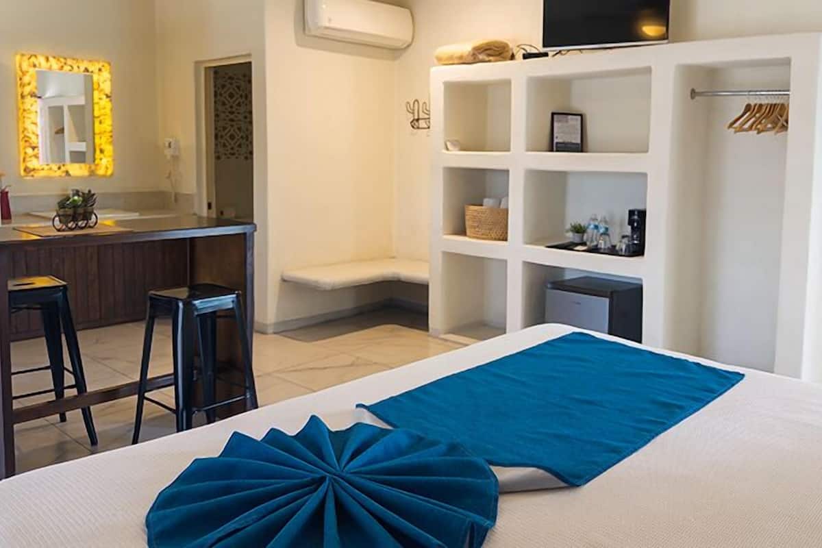 A hotel room with a bed, a desk with two chairs, a TV, a wardrobe, and a shelf with various items.