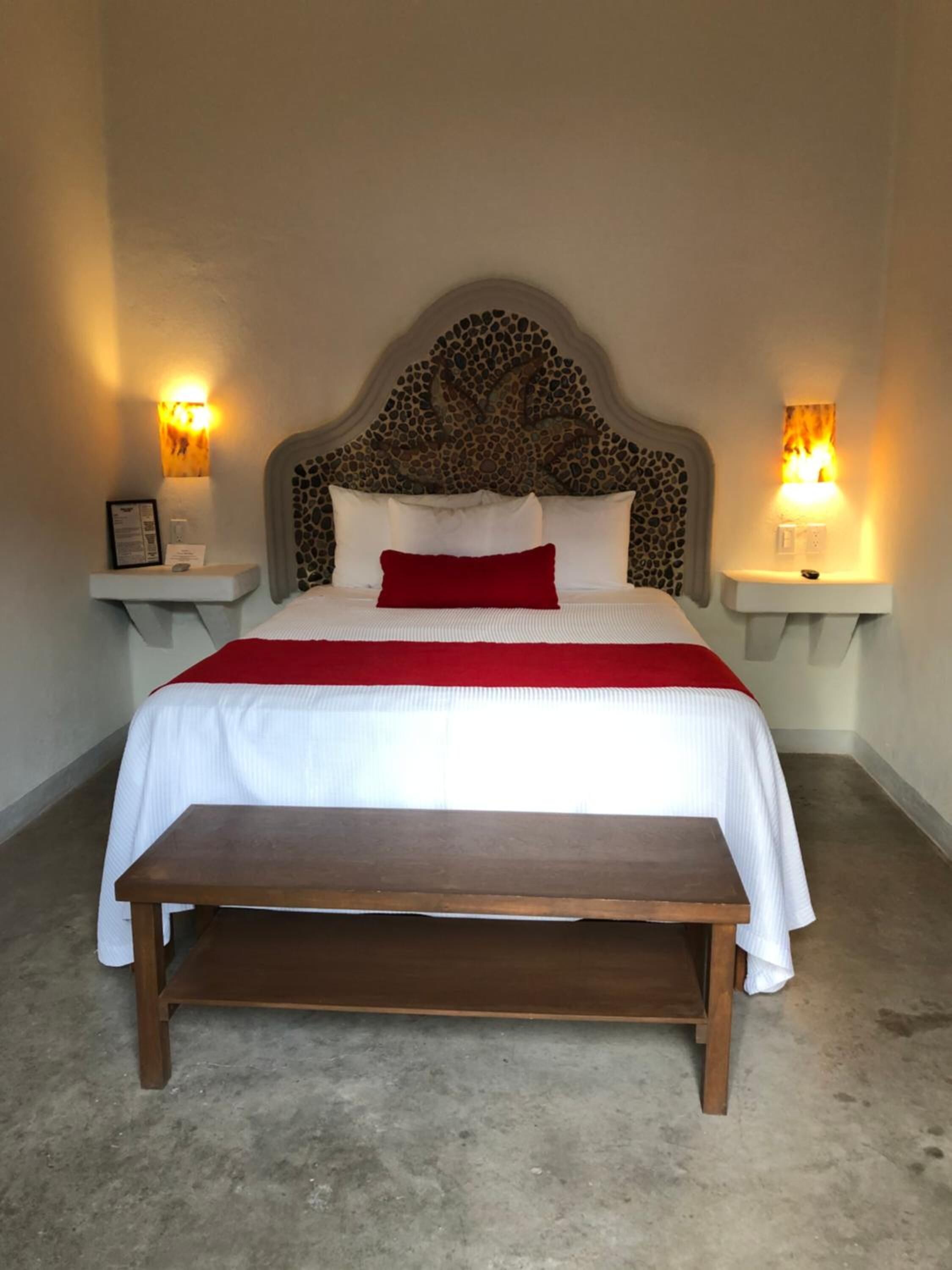 A bed with a stone headboard, white bedding, and red pillows. There are bedside tables with wall-mounted lamps. A wooden bench is in front of the bed.