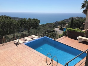 A swimming pool with a view of the sea, surrounded by greenery and a clear sky.