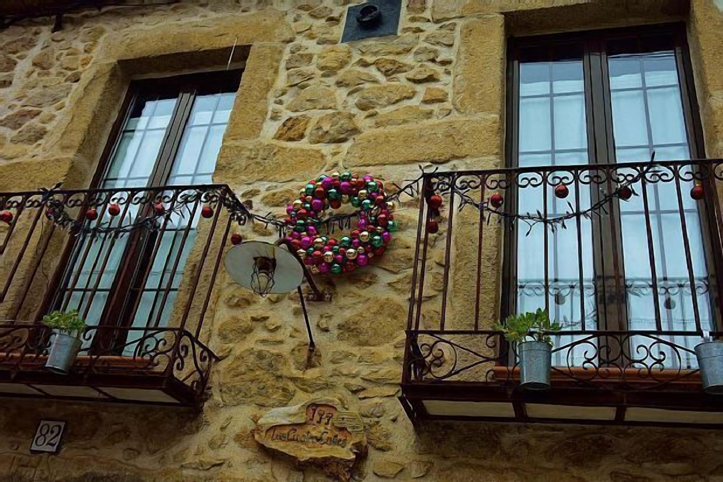A stone building with two windows, a decorative wreath, and potted plants on the balcony.