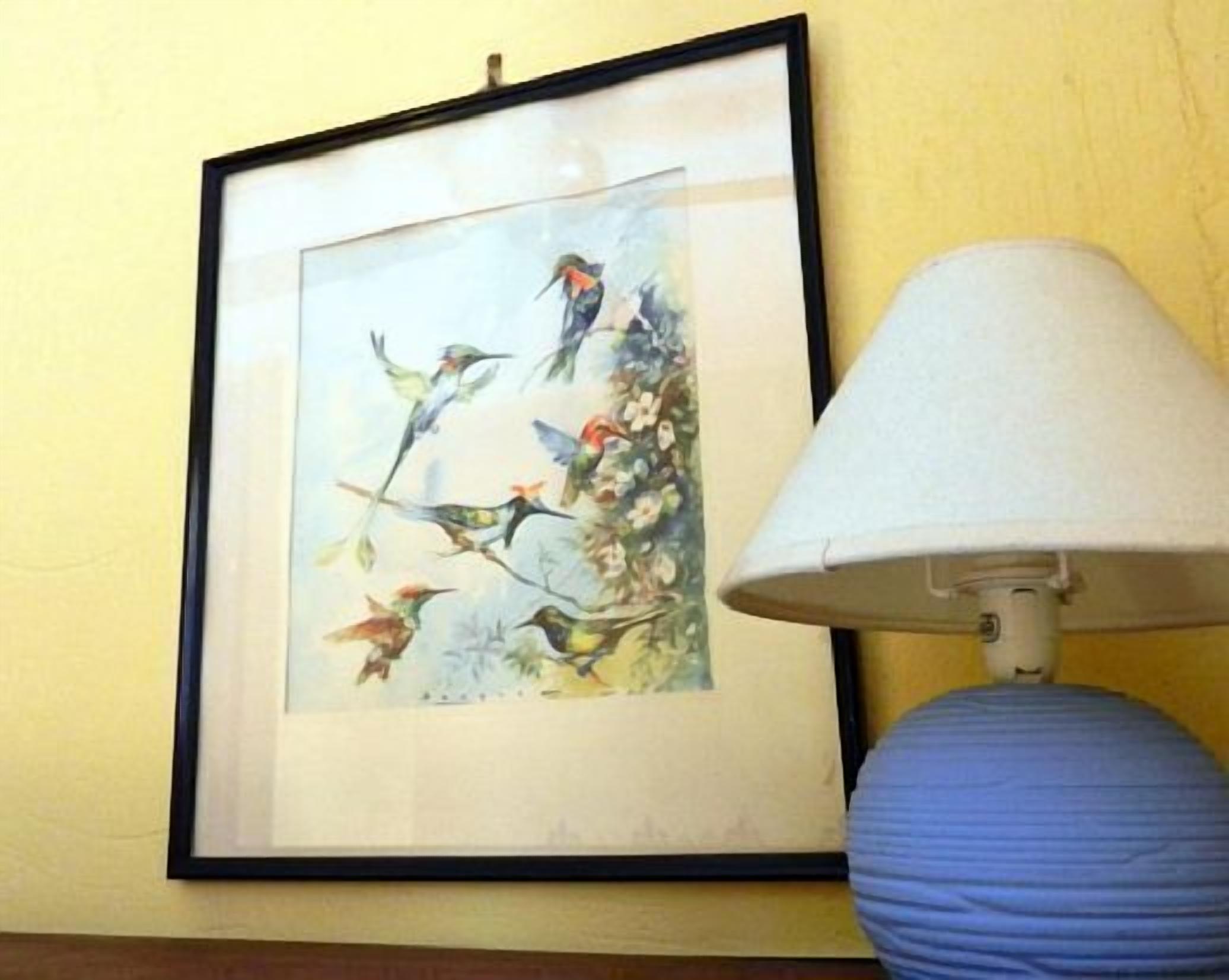 Uma pintura emoldurada com pássaros em um galho, um abajur branco e uma parede amarela.