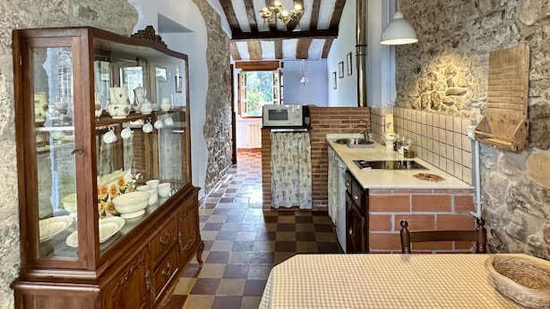 A rustic kitchen with exposed beams, a wooden cabinet with glass doors, a checkered tablecloth, and a hanging light fixture. - Rural Accommodations Casa Fabián, La Cabaña (Pet Friendly)  (Castrillón)