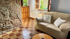 A cozy room with a checkered floor, a sofa with pillows, a wooden table with a remote control, and a stone wall. - Rural Accommodations Casa Fabián, La Cabaña (Pet Friendly)  (Castrillón)