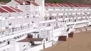 Un hôtel en bord de mer, avec plusieurs bâtiments blancs alignés, une plage de sable au premier plan et des falaises rocheuses en arrière-plan.