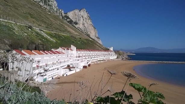 Un hôtel en bord de mer, avec une longue rangée de chambres donnant sur la plage, une falaise rocheuse et un ciel bleu dégagé.