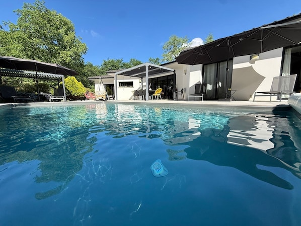 A clear swimming pool with lounge chairs and umbrellas, surrounded by a modern building with large windows and a covered patio area.