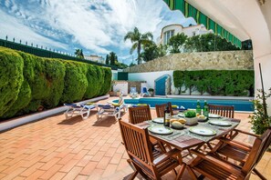 An outdoor patio with a pool, neatly arranged wooden chairs and tables, and a stone wall with greenery.