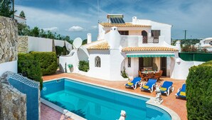 Una villa blanca con piscina, sillas de descanso azules y una estatua.