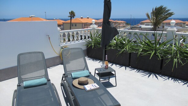 Terraza en la azotea con dos sillones, una mesa con una botella y un vaso, y vistas al mar y edificios.