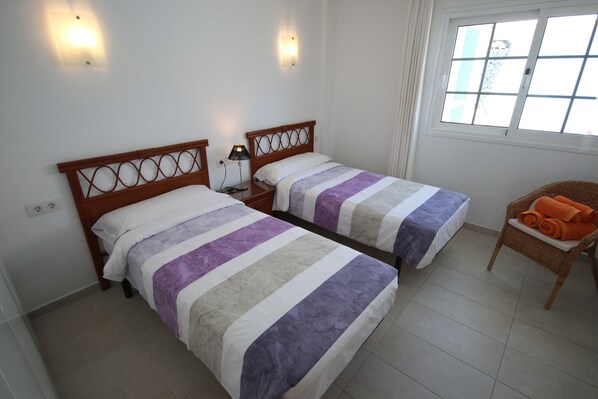 A hotel room with two beds, a nightstand, a chair with an orange towel, and a window with curtains. - LAS ROSAS by RENTMEDANO, stunning luxury Chocolate Box Villa (El Medano)