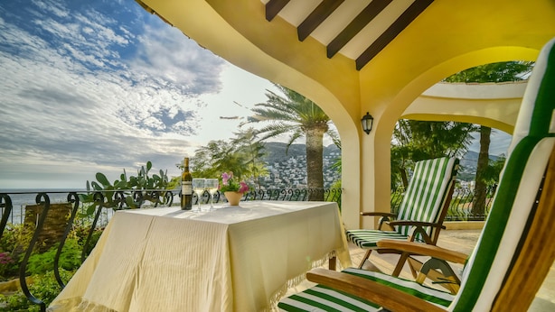 A covered outdoor seating area with a table and chairs, overlooking a scenic view of the sea and palm trees. - Family holiday house directly at sea (Calp)