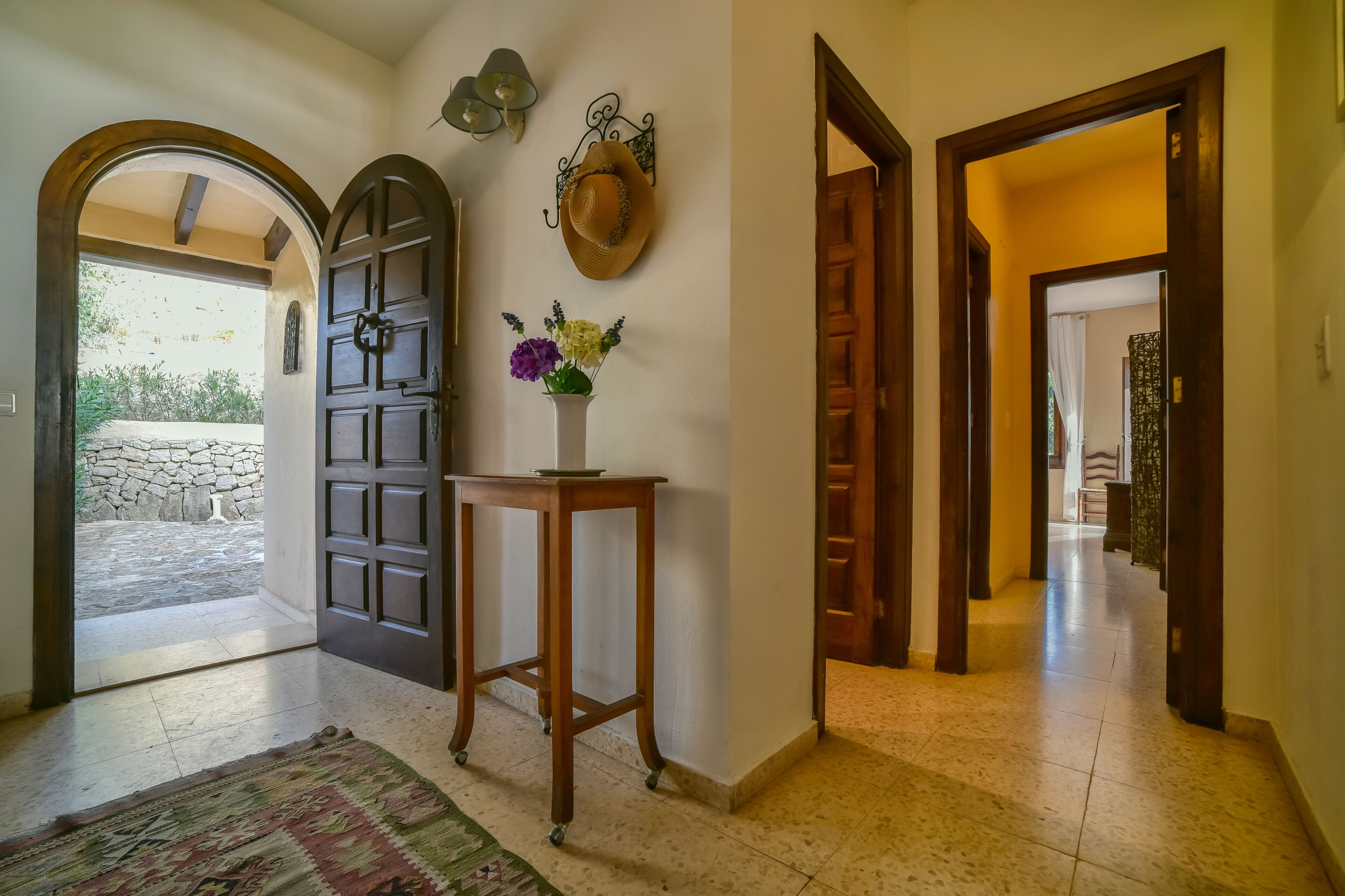 Un pasillo con una puerta de madera, una mesa pequeña con un florero y una pared de piedra visible a través de un arco.