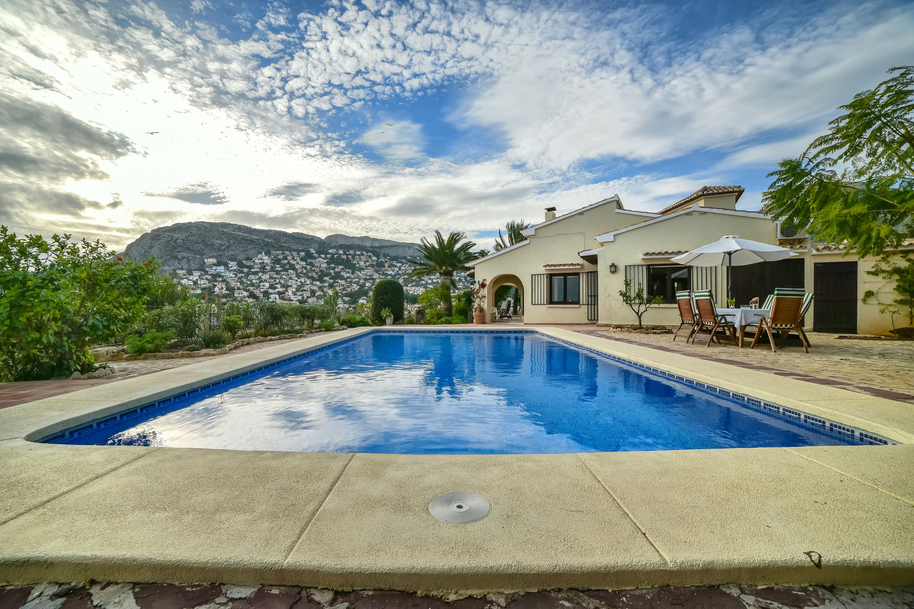 Une piscine avec un espace patio, un paysage montagneux en arrière-plan et une maison à la façade blanche.