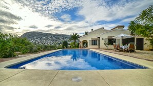 A swimming pool with a patio area, a mountain backdrop, and a house with a white facade. - Family holiday house directly at sea (Calp)