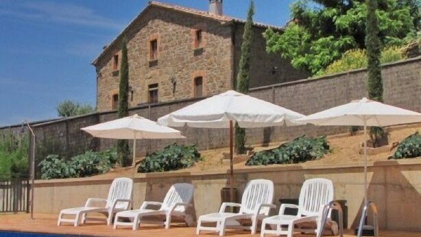 Un espace piscine avec des chaises longues blanches et des parasols, un bâtiment en pierre en arrière-plan et un ciel bleu dégagé.