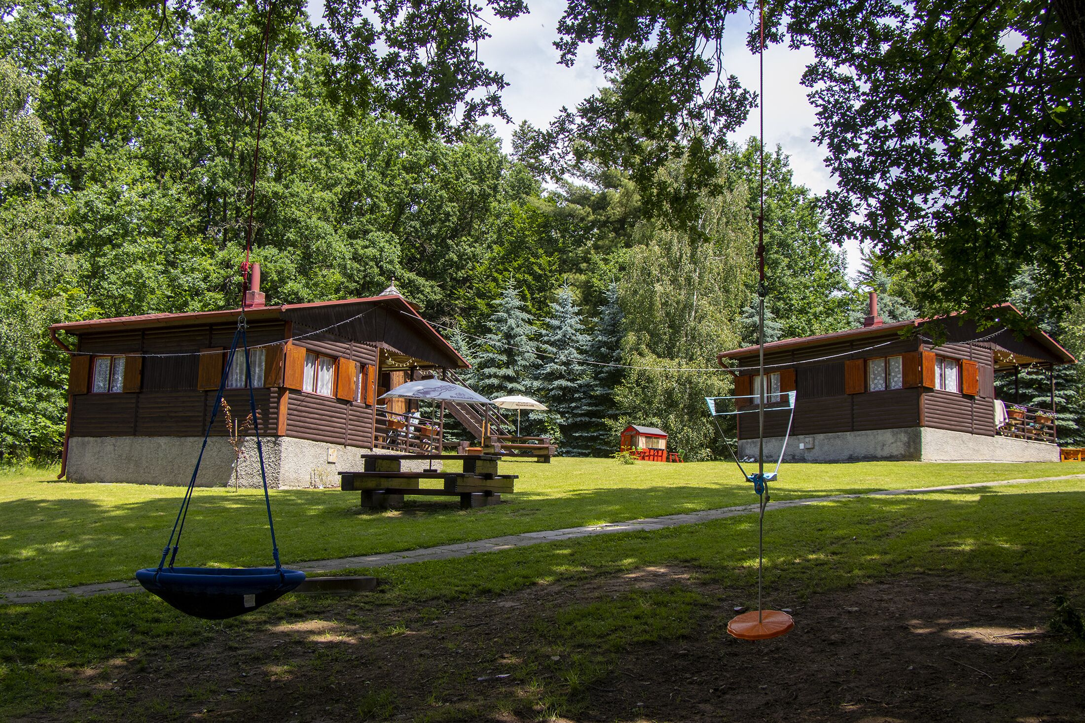 Two cabins with wooden exteriors, a swing set, picnic tables, and a small red shed in a wooded area.