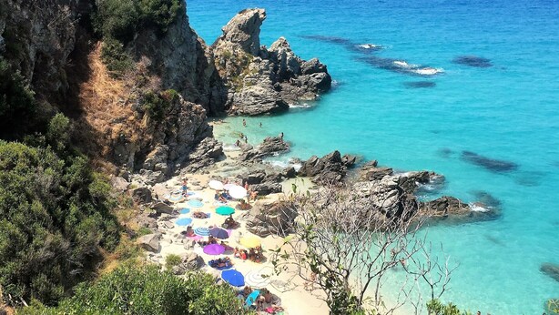 A coastal beach with clear turquoise waters, rocky cliffs, and a sandy area with colorful umbrellas. - New 4 bed villa in Zambrone - Spectacular 180 degree panoramic sea views. (Zambrone)