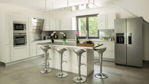 A modern kitchen with white cabinets, stainless steel appliances, a central island with bar stools, and a large window providing natural light.