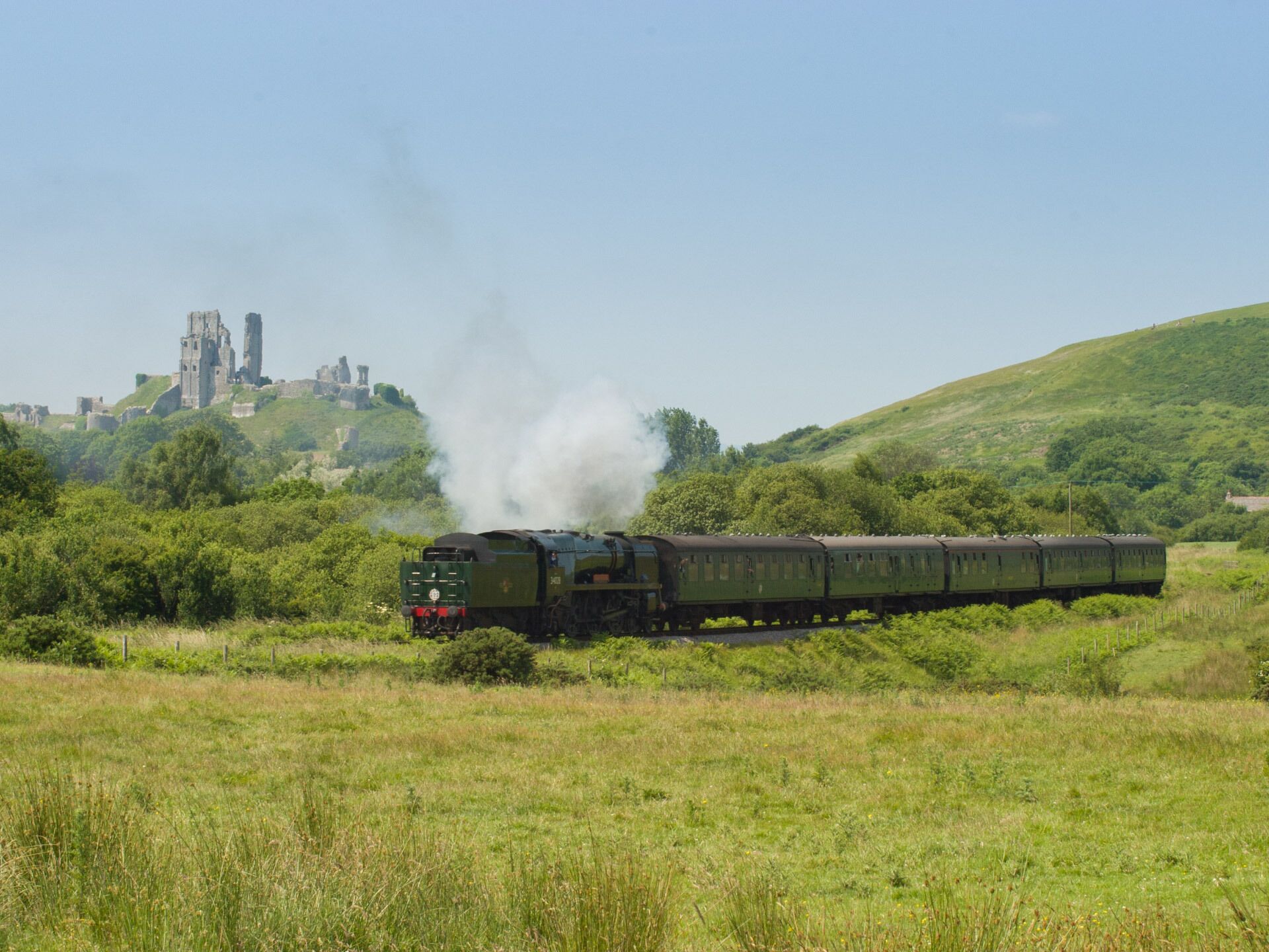 A steam locomotive with a series of passenger cars, traveling through a rural landscape with a castle in the background.