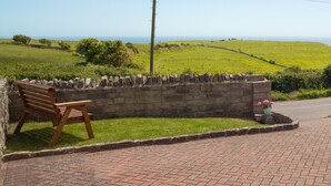Mural de pedra com um banco e um vaso de flores, com vista para um campo verde e o mar.