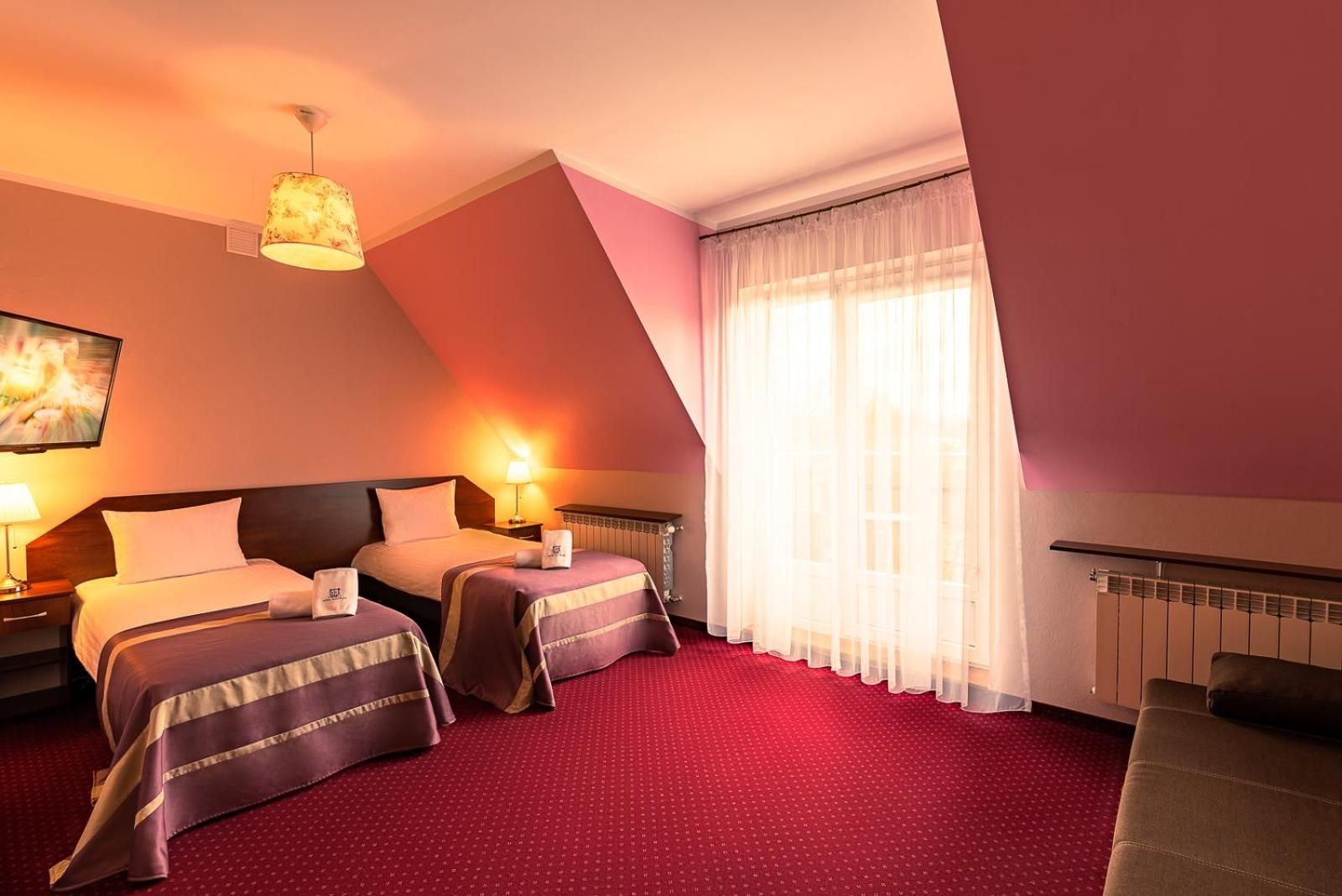 A hotel room with two beds, a red carpet, a large window with sheer curtains, and a ceiling light fixture.