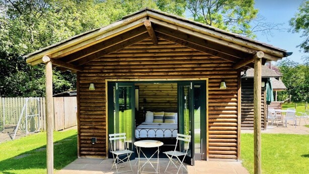 A wooden cabin with a covered patio, a bed, and two chairs.