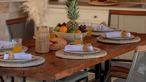Une table de salle à manger en bois, agrémentée de sets de table en fibres tressées, d’un vase contenant des plumes d’herbe des pampas, d’un saladier rempli de fruits, de croissants et de verres de jus d’orange.