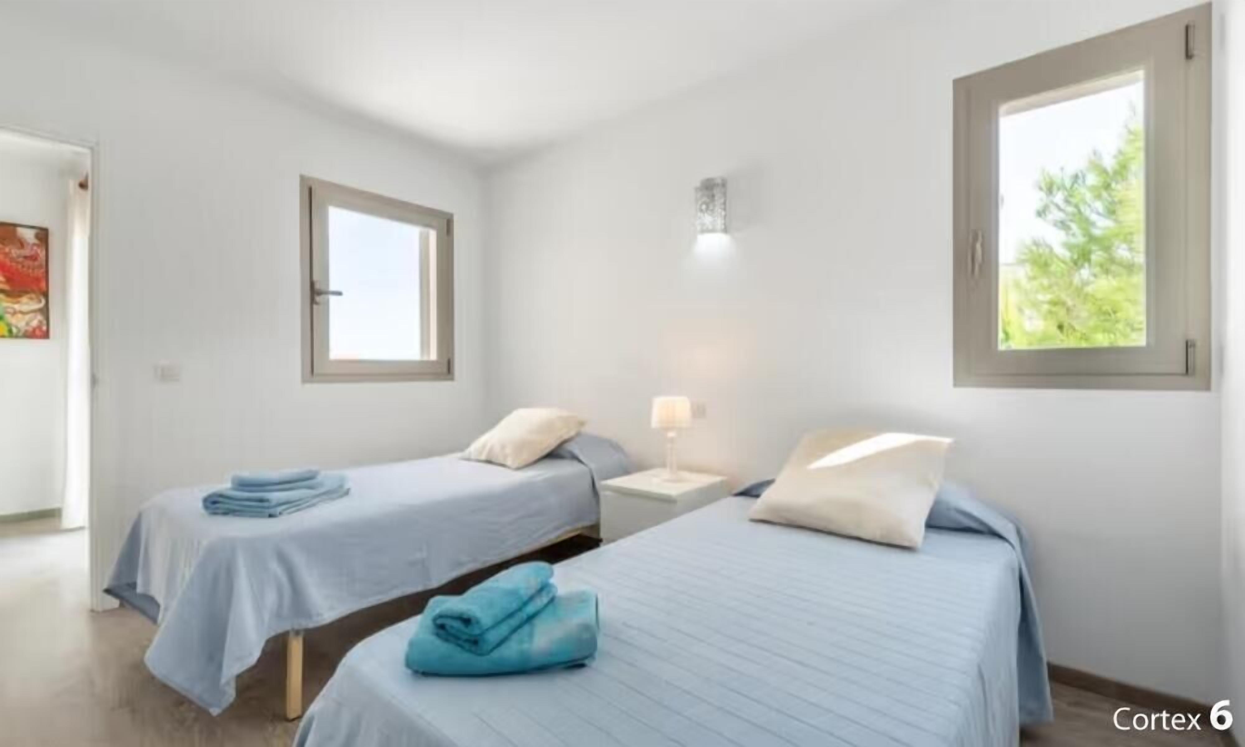 Une chambre avec deux lits, chacun recouvert d’une couverture bleue et agrémenté d’oreillers, une table de chevet avec une lampe, et une fenêtre donnant sur un paysage verdoyant.