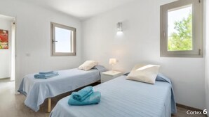 A room with two beds, each with a blue bedspread and pillows, a bedside table with a lamp, and a window with a view of greenery. - Large aloe house (Son Serra de Marina)