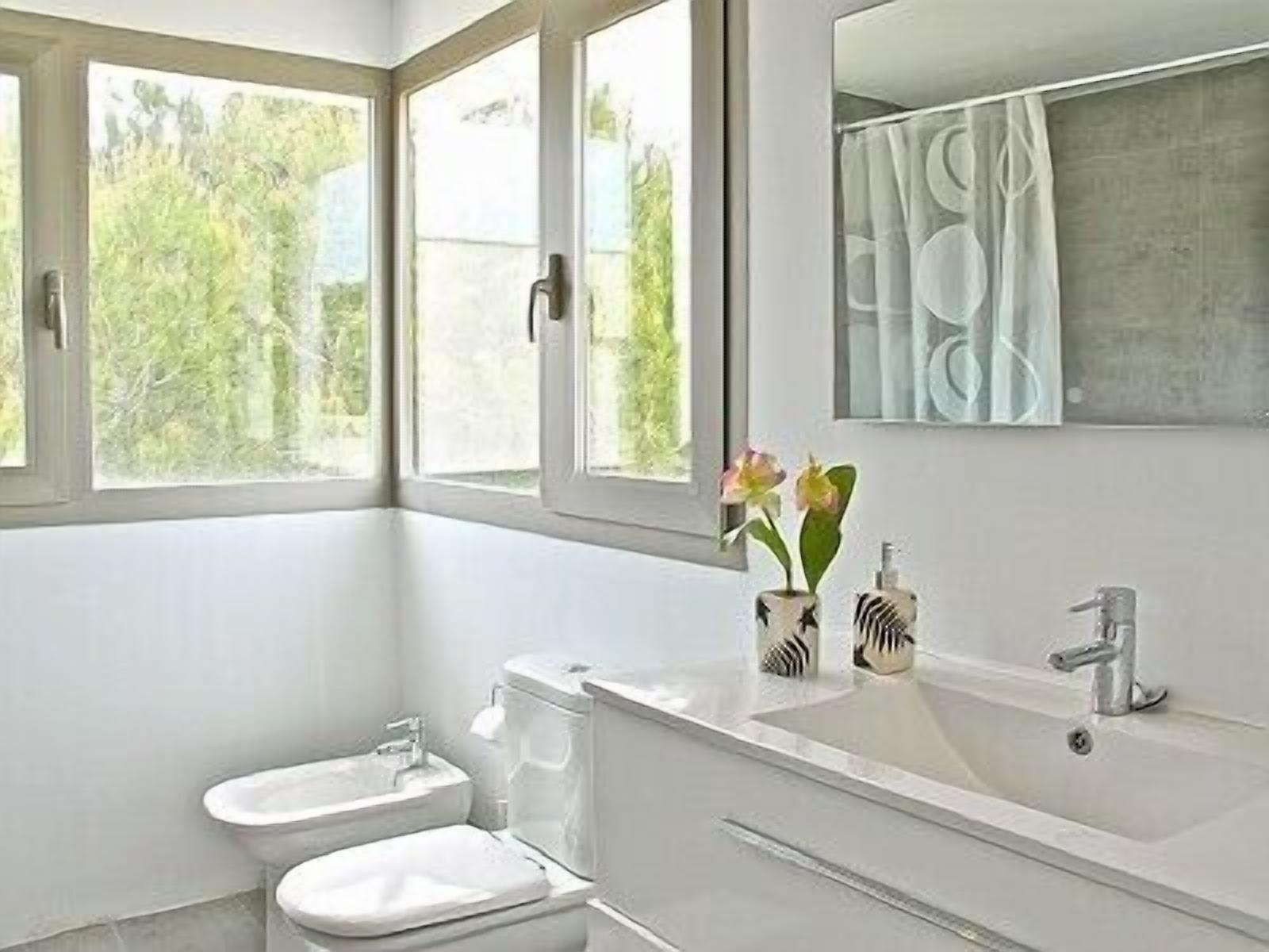 A modern bathroom with a toilet, sink, and bathtub. There are windows providing natural light and a view of greenery outside.