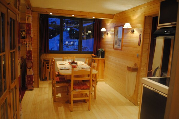 A wooden interior with a dining table set for four, a kitchen area with a refrigerator, and a window with a view of a snowy landscape.