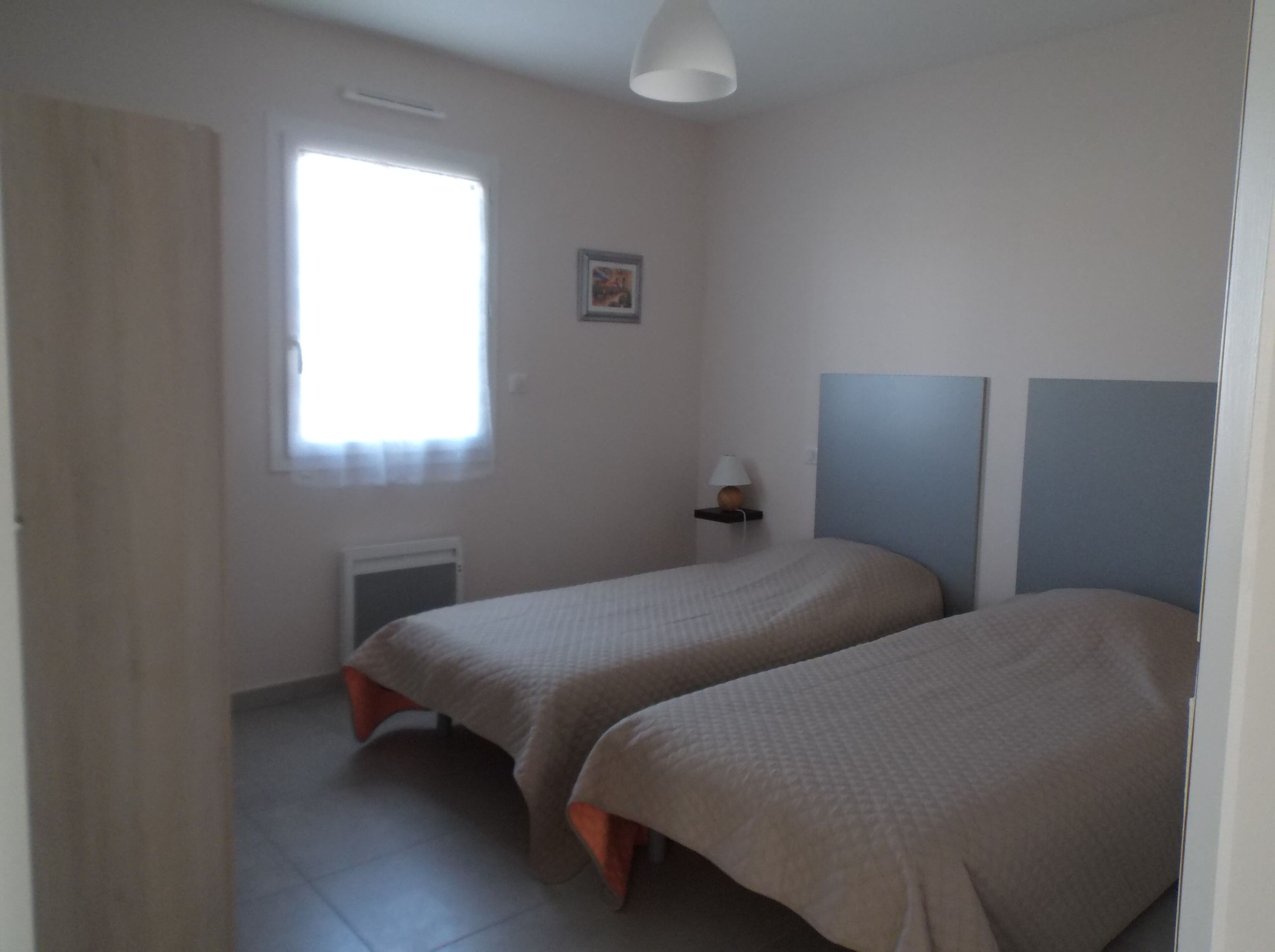 A bedroom with two beds, a window, a radiator, and a wall-mounted shelf with a lamp.