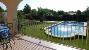 Une piscine entourée d’une clôture, un espace patio avec une table et des chaises, et une vue sur la verdure et les bâtiments au loin.