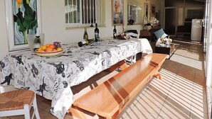 Un espace repas avec une longue table en bois recouverte d’une nappe à motifs, bordée de chaises blanches et d’un banc.