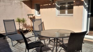 Een terras buiten met een ronde glazen tafel en vier rieten stoelen.