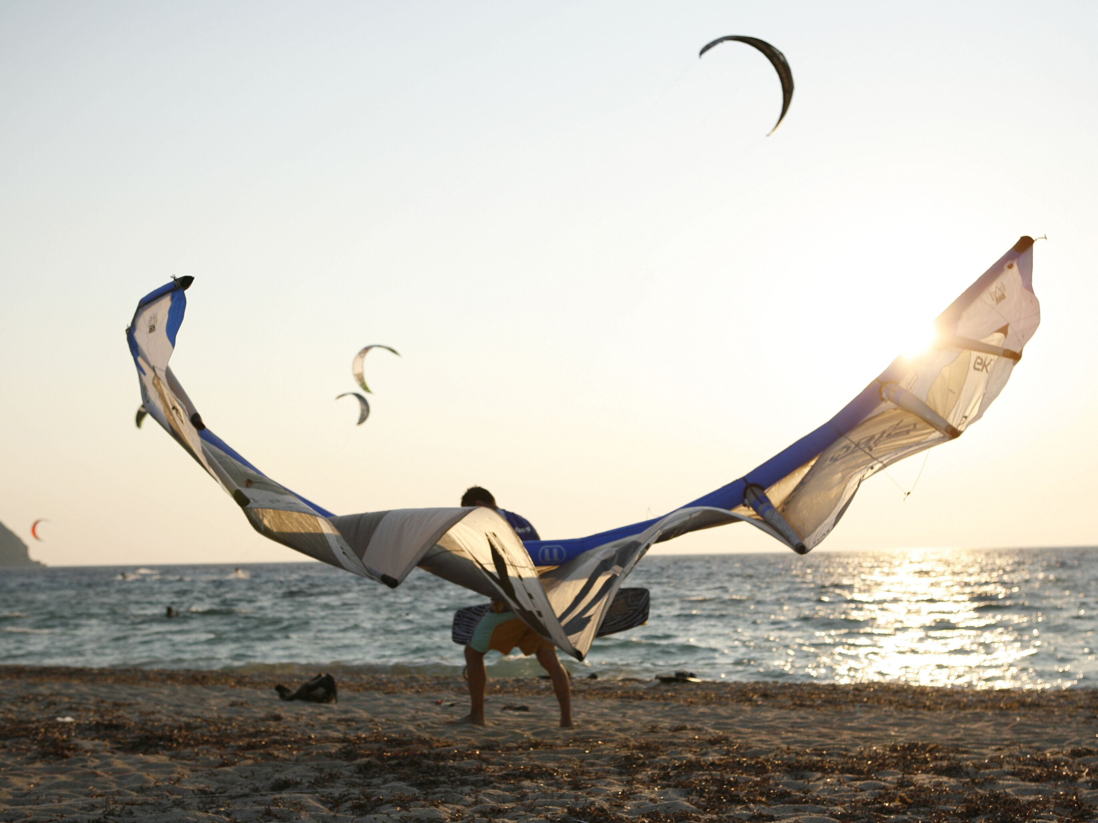Una playa con gente volando cometas y un velero a lo lejos.