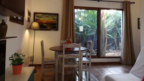 Ein Zimmer mit Esstisch, Stühlen, einem Blumentopf, einer Lampe, einem Gemälde und einer Glastür, die in einen Garten führt.