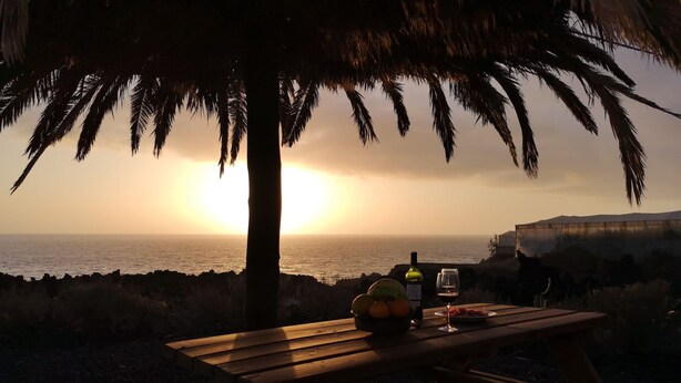 Ein Holztisch für ein Picknick mit einer Weinflasche, einem Glas und Obst unter einer Palme am Meer bei Sonnenuntergang.