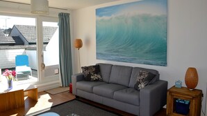 A living room with a grey sofa, a painting of a wave, and a view of a house through a glass door.