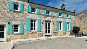 Un bâtiment en pierre doté de nombreuses fenêtres et d’une porte, avec une allée gravillonnée et un espace extérieur aménagé pour s’asseoir.