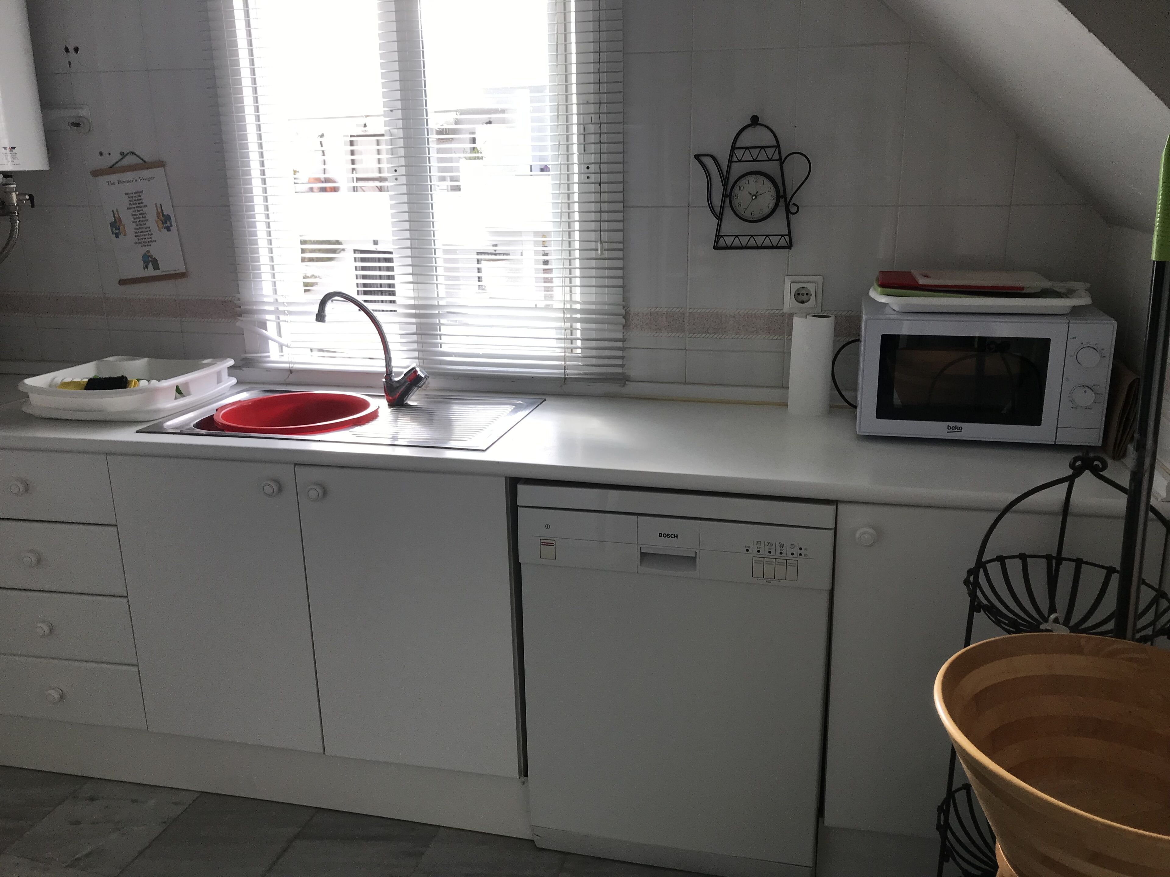 Una cucina con mobili bianchi, un lavandino rosso, una lavastoviglie Bosch, un forno a microonde e una finestra con persiane.
