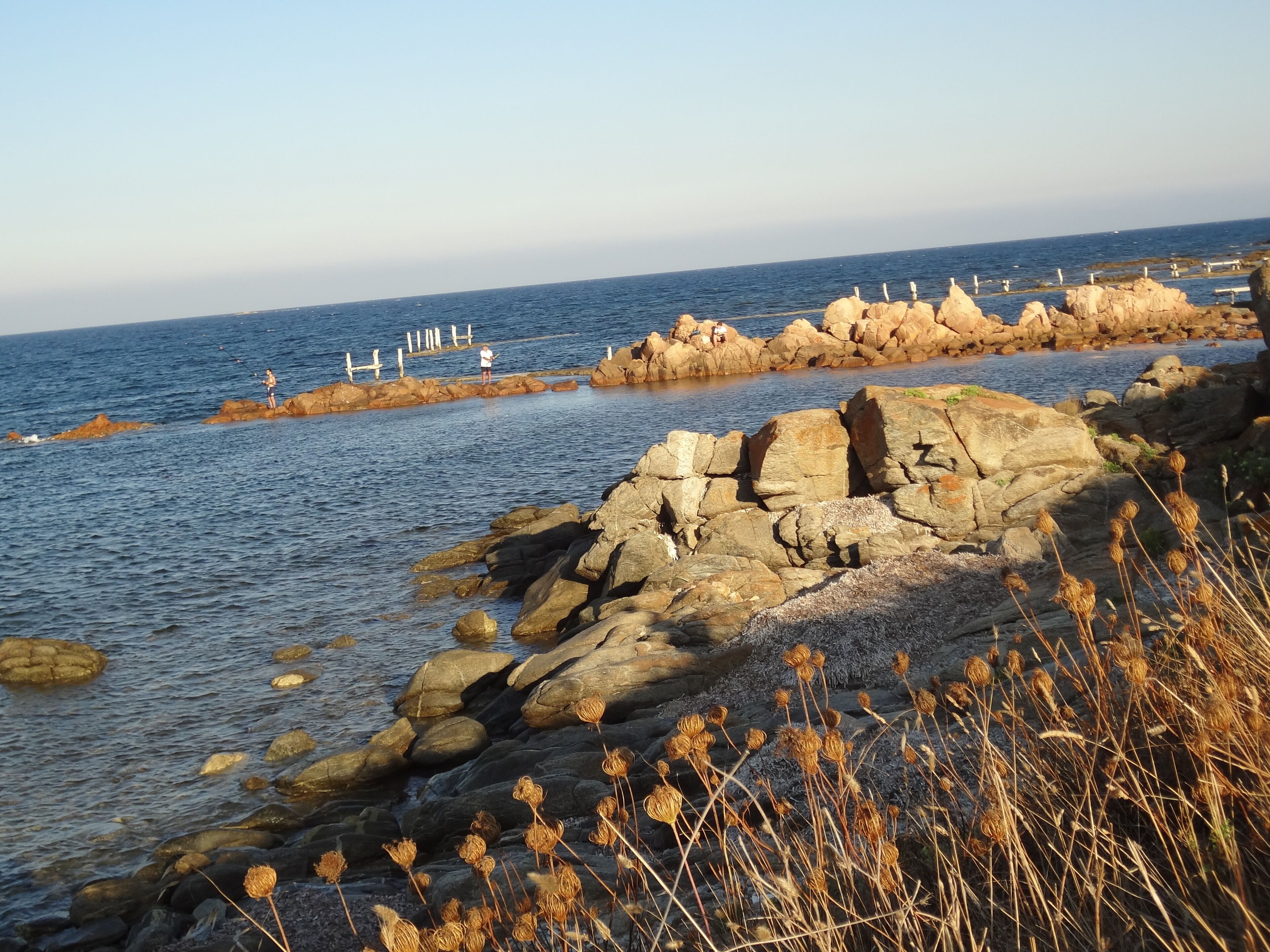 Eine felsige Küstenlinie mit freiem Blick auf das Meer und einer weit entfernten Felsenformation mit weißen Bauten.