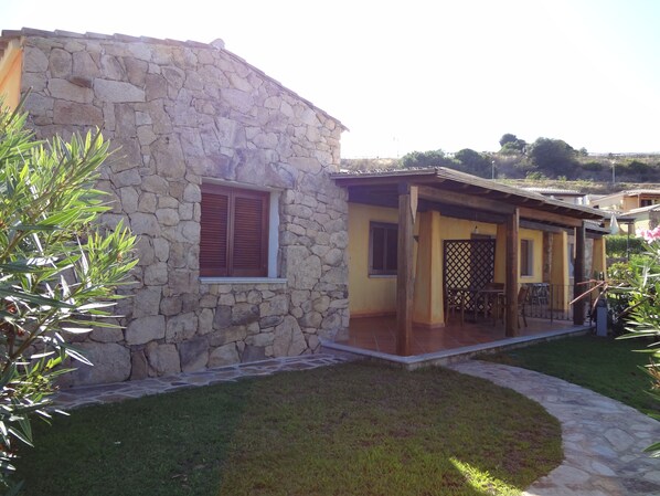 A stone house with a patio, a window with shutters, and a small lawn.