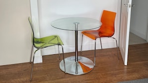 Et lille rum med et rundt glastilbord og to stole, en grøn og en orange, et ur på væggen og et indrammet billede over bordet.